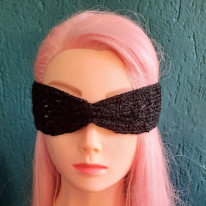 May include: A black, crocheted eye mask is displayed on a mannequin with pink hair. The eye mask is made of a textured yarn and covers the eyes. The background is a teal-colored wall.