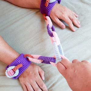 Könnte beinhalten: Ein Paar violette gehäkelte Handgelenksmanschetten, die durch eine mehrfarbige gehäkelte Kette verbunden sind. Die Kette besteht aus ineinandergreifenden Gliedern in Hellrosa, Hellblau und Violett. Jede Manschette hat ein gehäkeltes Schnallendetail.