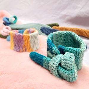 May include: A set of colorful crocheted headbands in shades of blue, green, yellow, and purple. The headbands are made with a variety of stitches and textures, and they are all tied in a knot at the back.