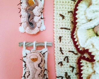 May include: Two crocheted wall hangings depicting the nativity scene. The larger hanging is cream with brown details and a white crocheted baby Jesus. The smaller hanging is white with a pink crocheted baby Jesus.