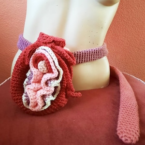 May include: A crocheted anatomical heart-shaped bag with a pink, red, and white color scheme. The bag is attached to a pink crocheted belt.