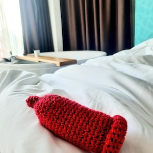 May include: A handmade, red crochet item, possibly a cozy or cover, rests on a white bed. The textured piece has a cylindrical shape with a rounded top and a flared base. The background includes a window, curtains, and a glimpse of a wooden tray.