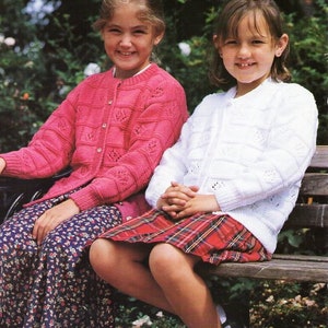 Może przedstawiać: Dwie dziewczyny w dzianinowych kardiganach, jeden różowy, a drugi biały, siedzą na drewnianej ławce. Dziewczyna w różowym kardiganie ma spódnicę w kwiaty, a dziewczyna w białym kardiganie ma spódnicę w czerwoną kratę.