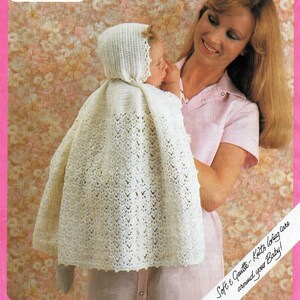 Può includere: Un berretto e una sciarpa per neonato in crochet bianco con un delicato motivo a pizzo. La sciarpa è drappeggiata su un bambino che una donna sta tenendo in braccio. Il testo "Soft & Gentle - Knits loving care around your Baby!" è scritto su un'etichetta bianca.