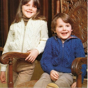 May include: Two children wearing knitted sweaters. The girl is wearing a white sweater with a cable knit pattern and a zipper. The boy is wearing a blue sweater with a cable knit pattern and a zipper. Both sweaters are sized 60-75 cm (24-30 inches).