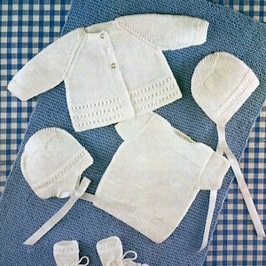 May include: A white knitted baby outfit set including a cardigan, a sweater, a hat, and booties. The set is laid out on a blue and white checkered fabric.