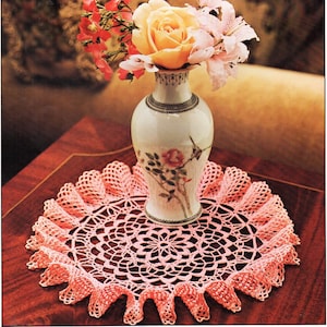 Puede incluir: Un encaje de ganchillo rosa con borde festoneado se encuentra sobre una mesa de madera. Un jarrón con diseño floral contiene un ramo de flores rosas y amarillas.