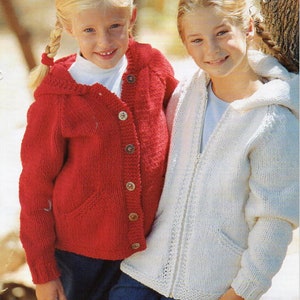 May include: Two young girls wearing knitted cardigans. The girl on the left is wearing a red cardigan with a hood and blue jeans. The girl on the right is wearing a white cardigan with a hood.