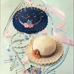 Pode incluir: Dois chapéus de crochê, um azul com um enfeite de flores e uma parte superior de almofada de alfinetes, o outro bege com um enfeite de flores. Ambos os chapéus são cercados por materiais de costura, incluindo alfinetes, botões e uma fita métrica.