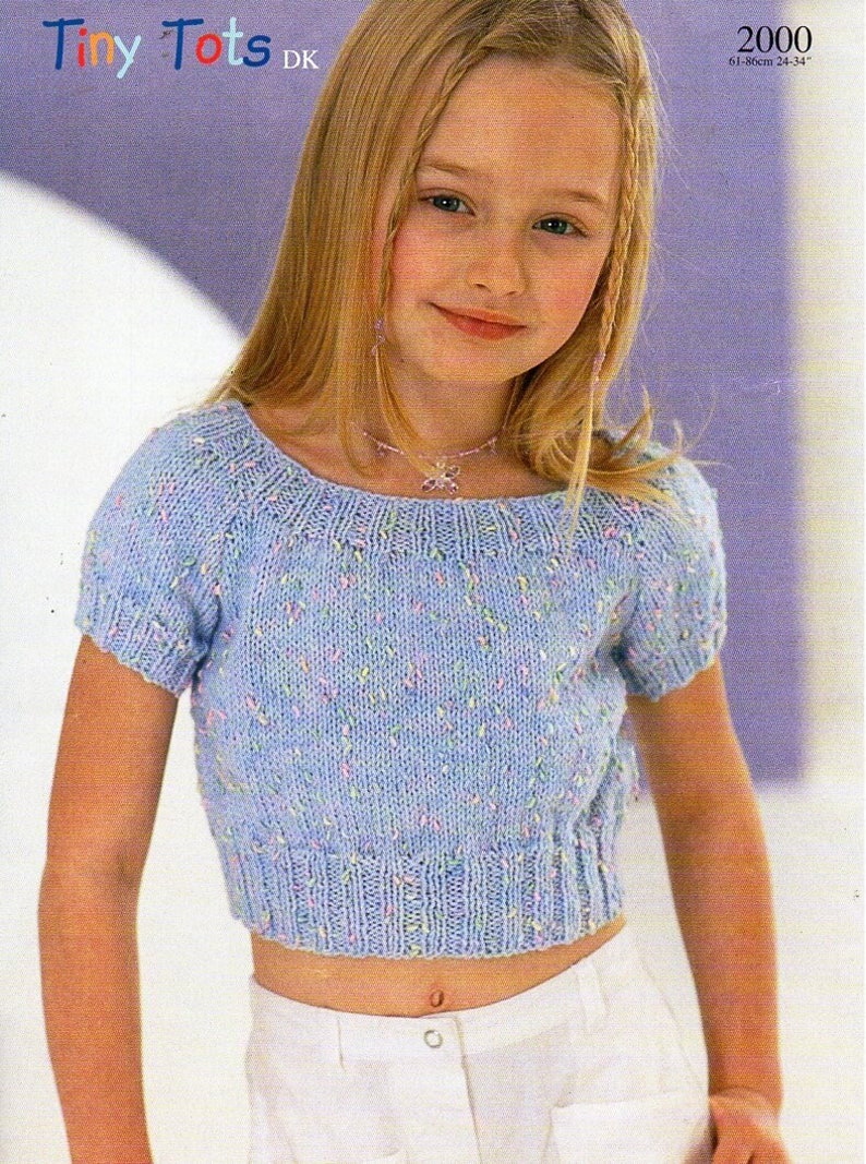 May include: A pale blue, short-sleeved knitted top with a round neckline. The top has a cropped design and is decorated with small, colourful flecks. The image also includes the text "Tiny Tots DK" and the numbers "2000".