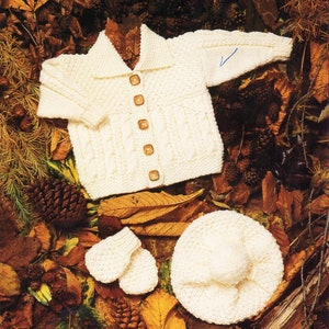 Puede incluir: Un conjunto de punto blanco para bebé compuesto por un cárdigan con botones, un gorro y mitones a juego. El conjunto está dispuesto sobre un lecho de hojas de otoño.