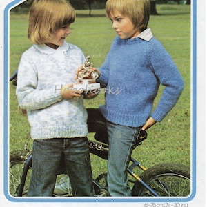 Pode incluir: Duas crianças vestindo suéteres de tricô, um azul e outro com um padrão branco e azul, estão ao lado de uma bicicleta. A criança com o suéter azul tem a mão no bolso. A criança com o suéter estampado está segurando um pequeno troféu.