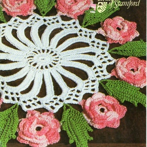 Könnte beinhalten: Eine weiße gehäkelte Tischdecke mit einem zentralen Blumendesign, umgeben von rosa und weißen gehäkelten Rosen und grünen Blättern. Die Tischdecke liegt auf einer dunkelbraunen Oberfläche. Das Bild enthält den Text "Twillleys of Stamford".