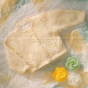 May include: A cream colored knitted baby cardigan with a delicate lace pattern. The cardigan is laid out on a textured background with three small yellow, white, and green rose-shaped candles.