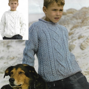 Puede incluir: Un suéter de punto de cable azul y blanco para niños. El suéter tiene un cuello redondo y mangas largas. El suéter se muestra en un niño que lleva jeans y está sentado junto a un perro.