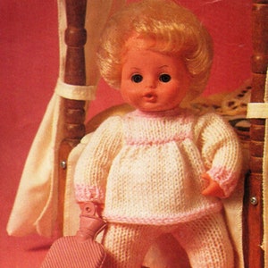 May include: A knitted doll outfit in pink and white, with a hot water bottle. The doll is wearing a long-sleeved sweater and pants. The outfit is made with a simple knit stitch.