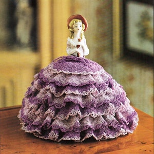 May include: A porcelain doll wearing a purple crocheted dress with multiple layers of ruffles. The doll is wearing a red hat and holding a small bouquet of flowers.