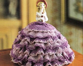 May include: A porcelain doll wearing a purple crocheted dress with multiple layers of ruffles. The doll is wearing a red hat and holding a small bouquet of flowers.