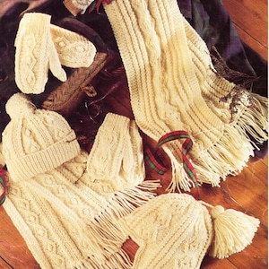 May include: Cream-colored knitted winter accessories set, including a hat with a pom-pom, a scarf with fringe, and mittens. The set features a cable knit design and is displayed on a wooden surface.