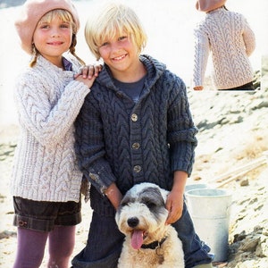 May include: Two children wearing knitted sweaters and a dog stand on a beach. The girl is wearing a pink beret and the boy is wearing a blue sweater with a hood. The dog is white with black markings. The text "BONUS ARAN 2332 4-13 years" is visible in the top right corner of the image.
