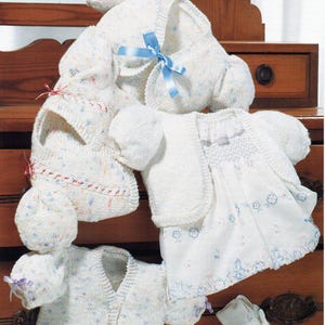 May include: A set of knitted baby clothes in white with a floral pattern. The set includes a cardigan, a hat, a pair of booties, and a dress.