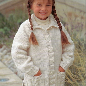 May include: A young girl wearing a white knitted cardigan with a button-up front and pockets. She has long brown hair in braids.