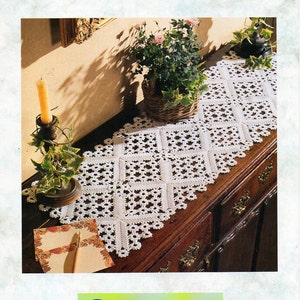 Op de afbeelding: Een witte gehaakte tafelloper met een vierkant patroon, gelegd op een houten tafel met een kandelaar en een potplant. De tafelloper is gemaakt met Coats Virtuoso garen. De afbeelding is afkomstig uit een Coats haakboekje, CCL 028.