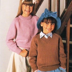 Puede incluir: Dos chicas con suéteres de punto. La chica de la izquierda lleva un suéter rosa y una falda blanca. La chica de la derecha lleva un suéter marrón y pantalones vaqueros azules con un parche de corazón rojo y blanco. Ambas chicas llevan sombreros.