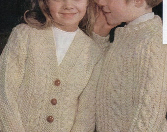 May include: Two children wearing cream-colored knitted sweaters. The girl is wearing a cardigan with brown buttons and a white undershirt. The boy is wearing a crew neck sweater. Both children are looking at each other.