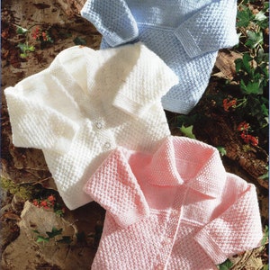 以下が含まれることがあります： 淡いパステルカラーの、水色、白、ピンクの3色のベビーニットセーター。長袖でボタン留めです。天然の木と葉の背景に配置されています。