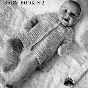 May include: A black and white photograph of a baby wearing a striped knitted outfit and a knitted hat. The baby is lying on a quilted blanket. The image includes the text "Quickerknit Baby Book N°2 9246-9° Designs for the first 6 months in Patons Quickerknit Baby Wool".