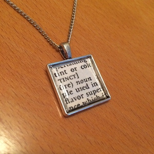 Pode incluir: Um colar de corrente de prata com um pingente de prata que apresenta a definição do dicionário da palavra "tinct" impressa em um fundo branco.