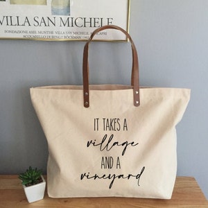 May include: A beige canvas tote bag with brown leather handles. The bag has a black graphic that reads "It takes a village and a vineyard".