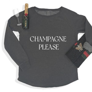 May include: A dark gray long-sleeved sweatshirt with the text "CHAMPAGNE PLEASE" in white. A bottle of Moët champagne and a champagne glass are to the left of the shirt. A black clutch with a bee and red and green stripes is to the right.