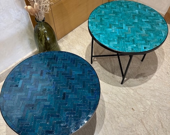 Op de afbeelding: Twee ronde tafels met een mozaïek tegelblad en zwarte metalen poten. De tafels staan op een lichtbeige betegelde vloer. Het blad van de tafel links is donkerder blauw dan de tafel rechts.