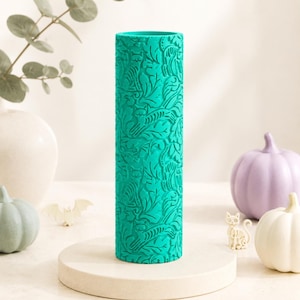 May include: A cylindrical teal rolling pin featuring an embossed pattern of cats and leaves. The surface is textured with repeating feline figures and foliage motifs, designed for creating impressions in clay or dough, standing upright on a base.
