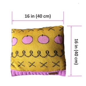 May include: A square, knitted pillow, 16 inches (40 cm) on each side, with a mustard yellow background. The pillow features pink fruit designs, black cross and squiggle patterns, and a pink border. A small label is visible.