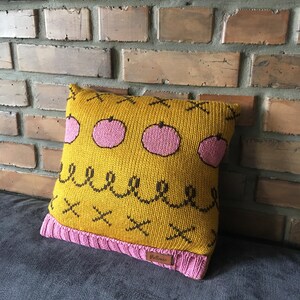 May include: A square knitted pillow with a mustard yellow background and pink apple designs. The pillow has a pink knitted border and a brown label. The design includes black squiggles and X's.