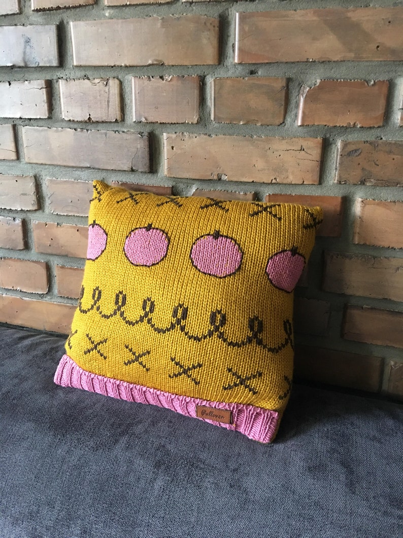 May include: A square knitted pillow with a mustard yellow background and pink apple designs. The pillow has a pink knitted border and a brown label. The design includes black squiggles and X's.