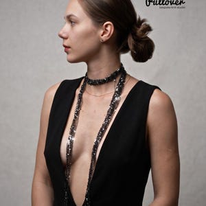 May include: A long, black, shimmering beaded necklace featuring multiple strands of dark, reflective sequins and beads that drape down the chest. The necklace is worn with a sleeveless black dress with a deep V-neckline. Text reads Pullover bespoke knit studio.