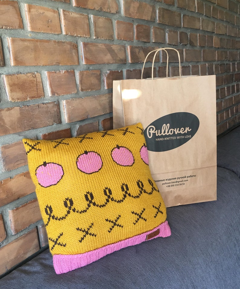 May include: A square, mustard-yellow knitted pillow with pink accents and a decorative pattern of pink circles, black squiggles, and black X's. The pillow has a pink border and is next to a brown paper shopping bag with the word "Pullover" on it.
