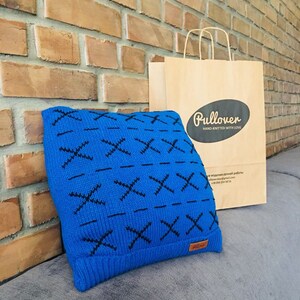 May include: A vibrant blue knit hat with a black cross-stitch pattern. The hat is displayed next to a brown paper shopping bag with the word "Pullover" and the phrase "Hand-Knitted With Love" printed on it. The background features a brick wall.