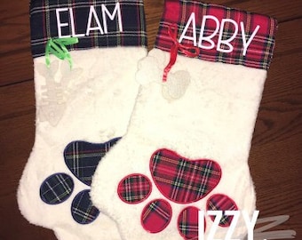 May include: Two white Christmas stockings with red and blue plaid paw prints. The stockings have red and blue plaid cuffs with the names "Elam" and "Abby" embroidered on them.