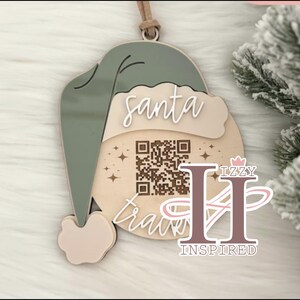 May include: A Christmas ornament shaped like Santa's hat, in shades of green, beige, and light pink. The ornament has the words "santa travels" and a QR code. The ornament is made of wood and has a leather string for hanging.