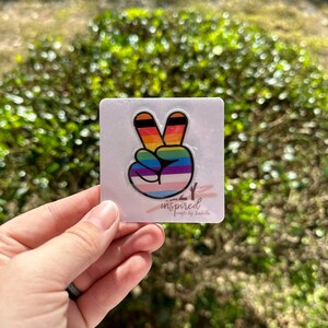May include: A rainbow peace sign sticker with black outline. The sticker is on a white background with the text "inspired crafts by Jaquelle" in black.