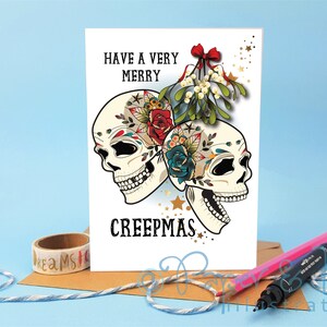 May include: A white greeting card with a festive message that reads "Have a Very Merry Creepmas". The card features two colorful sugar skulls with floral designs and a sprig of mistletoe with a red bow.
