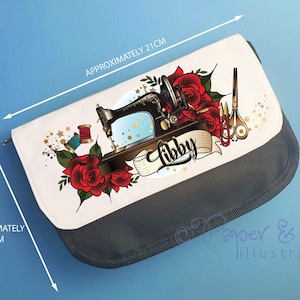Puede incluir: Un estuche de lápices blanco con cremallera y ribete negro. El estuche presenta una máquina de coser vintage con rosas rojas, estrellas doradas y el nombre "Libby" en letras doradas. El estuche mide aproximadamente 18 cm por 36 cm.