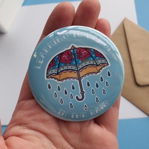 May include: A blue round button with a white outline and the text "Learning to Dance in the Rain". The button features a colourful illustration of an umbrella with red roses and blue and yellow accents. Raindrops are falling from the umbrella.
