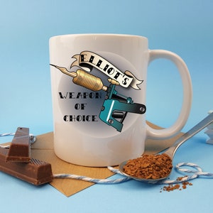 May include: White ceramic mug with a tattoo machine design and the text "Elliot's Weapon of Choice".