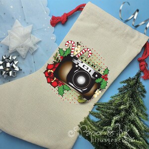 Op de afbeelding: Een beige kerstsok met een rood en groen hulstontwerp en een vintage camera met de tekst "Let It Roll".